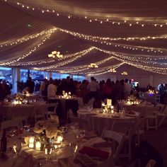 a room filled with lots of tables covered in white linens and lit up by string lights