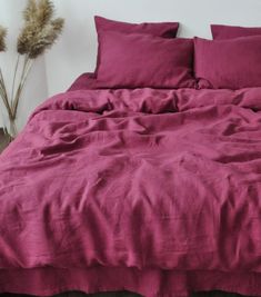 a bed with pink sheets and pillows in a white room next to two dried plants