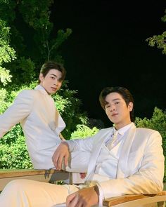 two men in white suits sitting on wooden chairs with trees in the background at night