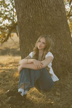 a woman sitting under a tree with her legs crossed in front of her face and looking at the camera