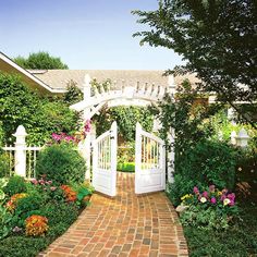 a brick pathway leads to an open white gated garden area with flowers and greenery