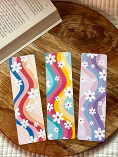 three colorful bookmarks sitting on top of a wooden table next to an open book