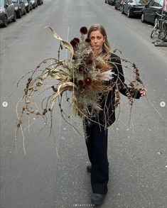 a woman is walking down the street with dried plants in her hands and flowers on her shoulder