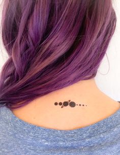 a woman with purple hair has her back turned to the camera and is wearing a small black dot tattoo on her neck