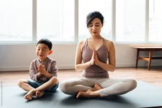 a woman and child sitting on yoga mats in front of large windows, meditating