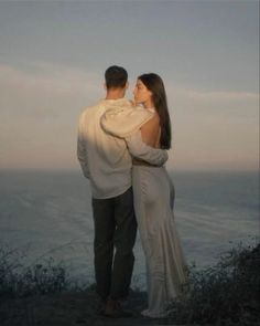 a man and woman standing next to each other on top of a hill near the ocean