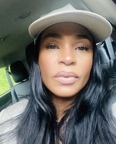 a woman with long black hair wearing a baseball cap and looking at the camera while sitting in a car