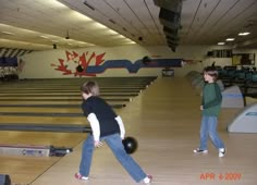 two young boys playing bowling in an indoor bowling alley with balls on the floor and lanes lined up behind them