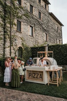 a group of people standing around a pizza stand in front of a stone building with ivy growing on it