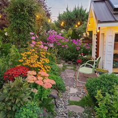 a garden with lots of flowers next to a small yellow building and lights hanging from the roof