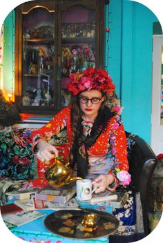 a woman sitting at a table in front of a tea pot and cup with flowers on her head