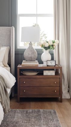 a bedroom with a bed, nightstand and flowers on the night stand in front of the window