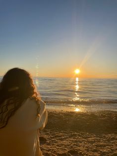 a woman is sitting on the beach watching the sun go down over the ocean with her hair blowing in the wind