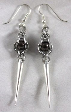 This product of Cassie's Creations is made from silver anodized aluminum rings that cage in a brown pearl bead, with silver spike charms that dangle down. The earrings are hung on black earring hooks. To get a list of the shows where we will be, visit www.MoonDragonDesigns.com, or message for custom orders Beaded Earrings Ideas, Chain Mail Earrings, Chainmaille Jewelry Patterns, Black Earring, Jump Ring Jewelry, Chainmail Jewelry, Chain Maille Jewelry, Bead Weaving Patterns, Chain Maille