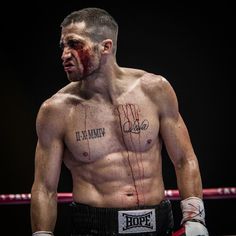 a shirtless man with blood on his face standing in a ring wearing boxing gloves