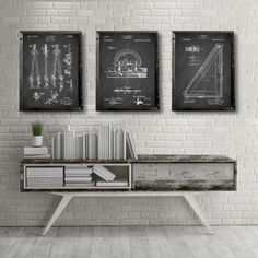 three black and white prints on a brick wall above a desk with books, plant and pencils