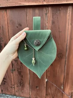 a person holding up a green leaf shaped pouch with charms attached to it, in front of a wooden wall