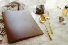 a table topped with a brown leather notebook and gold utensils next to a bottle of whiskey