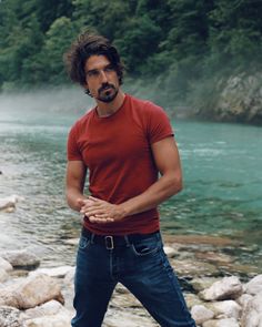 a man standing in front of a river with rocks on the ground and water behind him