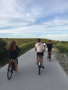 three people riding bikes down a paved road with grass and bushes in the background,