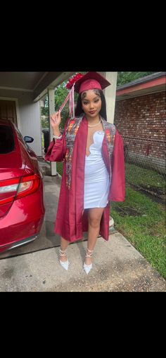 a woman standing in front of a red car wearing a graduation gown and holding her hand up