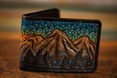 Discover the exceptional quality of our Men's Leather Wallet, featuring a unique sunset and mountain scene embellished with sacred geometry. Crafted from high-quality vegetable-tanned leather, this wallet showcases a colorful, hand-tooled design available in your choice of color scheme. Each wallet is meticulously dyed, stitched, and finished with a protective coating to ensure the leather and dye work remains vibrant and durable over time. Designed to be both practical and stylish, this wallet Handcrafted Leather Wallet, Tooled Leather Wallet, Mountains And Trees, Leather Craft Projects, Mens Tools, Brown Wallet, Mountain Designs, Blue Color Schemes, Mountain Scene