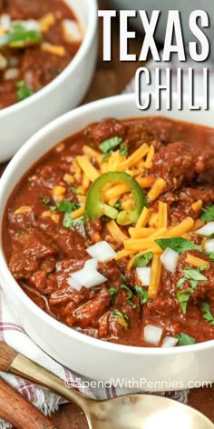 two white bowls filled with chili and cheese on top of a wooden table next to spoons