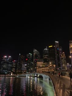 people are walking along the water in front of tall buildings at night time with lights reflecting on the water