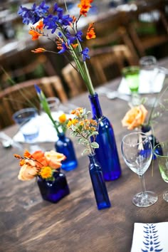 the table is set with blue glass vases and flowers in them, along with empty wine glasses