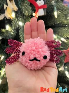 a hand holding a small pink pom - pom in front of a christmas tree