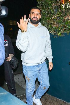 a man in grey sweatshirt and ripped jeans holding his hand up to the camera while standing outside