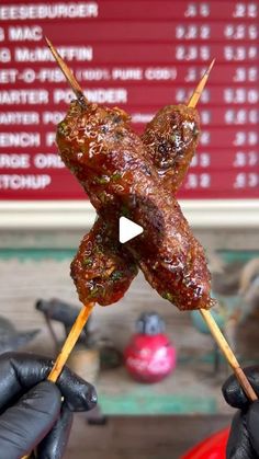 a person holding up two skewers with meat on them in front of a sign