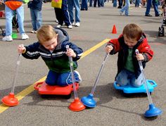 two young boys playing on small toy cars in an open area with people walking around