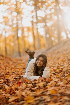 a woman laying on the ground surrounded by leaves in an autumn forest with sunlight shining through the trees