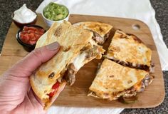 a person is holding up a sandwich on a cutting board with salsa and guacamole in the background