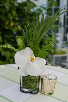 a white flower in a glass vase next to a metal cup on a checkered table cloth