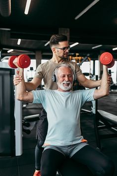 an older man doing exercises with dumbbells in the gym while another man watches