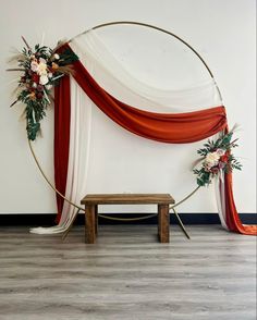 an arch decorated with red and white draping, flowers and greenery sits in front of a wooden bench