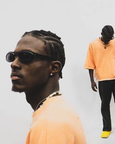 a man with dreadlocks standing in front of a white wall wearing an orange shirt and black pants