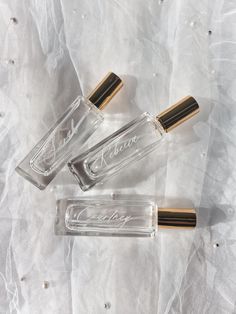 three clear bottles with gold caps sitting on a white sheeted surface, one has the word's name written in cursive writing