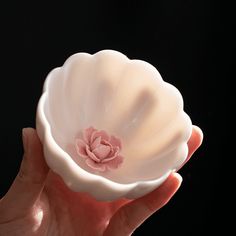 a hand holding a white bowl with a pink flower in it's center on a black background