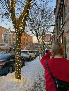 a woman walking down a snow covered sidewalk next to a tree with christmas lights on it