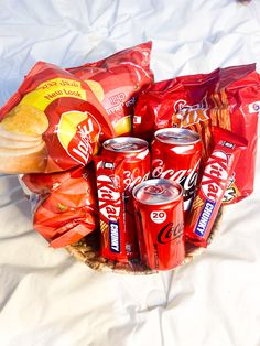 a basket filled with chips and sodas on top of a white sheet covered bed