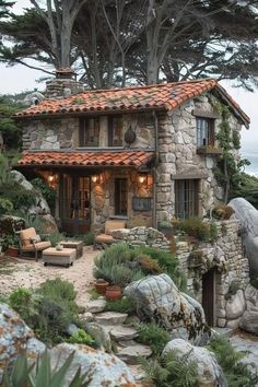 a small stone house surrounded by trees and rocks near the ocean with an orange tiled roof