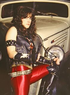 a woman in red pants and leather boots standing next to an old car with her hand on the steering wheel