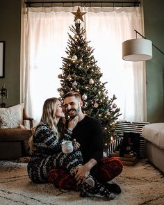 a man and woman are sitting in front of a christmas tree while holding coffee cups