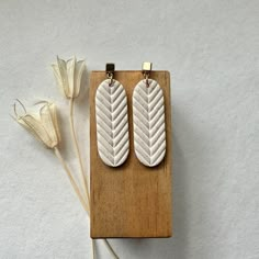 two white leather earrings sitting on top of a wooden board next to a dried flower