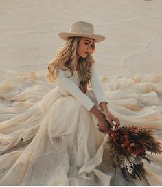 a woman in a white dress and hat sitting on the ground holding a dried plant