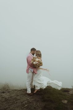 a bride and groom kissing on top of a hill in the foggy weather with their bouquet