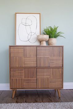 a wooden cabinet sitting next to a plant on top of a rug in front of a painting